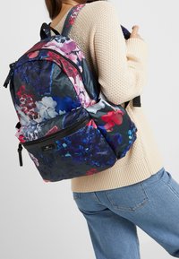 Henkilö, joka käyttää vaaleabeigeä neulepaitaa ja sinisiä farkkuja, kantaa suurta kukkakuvioista reppua, jossa on useita vetoketjullisia lokeroita.