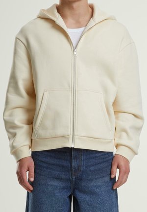 Person trägt einen cremefarbenen Hoodie mit Reißverschluss und Vorderentaschen sowie blaue Jeans, steht vor einem einfarbigen hellen Hintergrund.