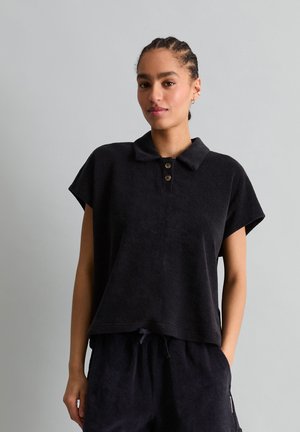 Kobieta z warkoczami, ubrana w luźną czarną koszulkę polo z krótkim rękawem oraz pasujące czarne spodnie ze ściągaczem, na jasnym jednolitym tle.