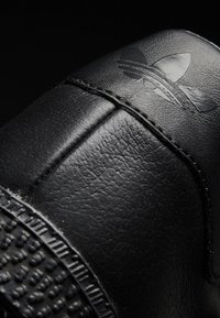 Zapatilla negra de cuero con paneles texturizados, que presenta un logo en relieve y una suela de goma estriada. La vista en primer plano resalta los detalles de la costura.