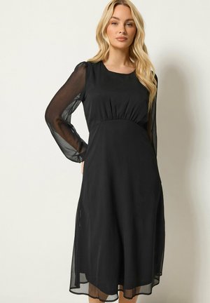 Blonde Frau trägt ein schwarzes Midikleid mit transparenten langen Ärmeln und steht vor einem einfarbigen hellen Hintergrund.