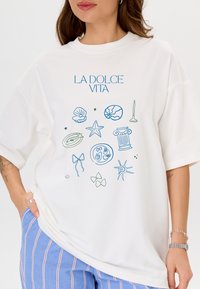 Witte oversized t-shirt met een blauw en groene grafische afbeelding met verschillende iconen en de tekst "LA DOLCE VITA" prominent weergegeven.