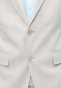 Veste de costume gris clair à deux boutons fermés sur une chemise bleue claire, montrant les détails avant et les poches.