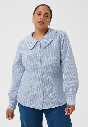 Femme portant une blouse à carreaux bleus et blancs avec des boutons, un large col et de longues manches bouffantes, associée à un jean bleu.