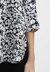 Svart och vit mönstrad blus med korta ärmar, med en lös passform och rundad nederkant. Tyget verkar lätt och svävande.