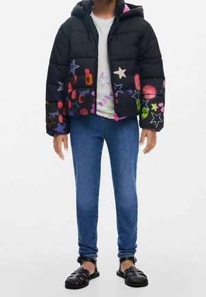 Enfant portant une veste matelassée noire avec des imprimés colorés d'étoiles et de formes, un jean bleu et des sandales noires sur fond blanc.