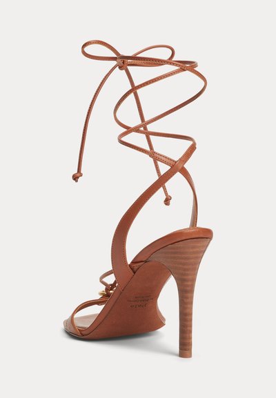 Καφέ σατινέ δερμάτινο σανδάλι με ψηλό τακούνι και λεπτά λουριά που τυλίγονται γύρω από τον αστράγαλο σε στυλ δεσίματος με κορδόνια, σε λευκό φόντο.