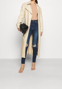 Mujer vistiendo un abrigo largo beige sobre una blusa color arena y jeans rotos azules, sosteniendo un clutch negro, de pie en sandalias de tacón color arena, contra un fondo blanco.