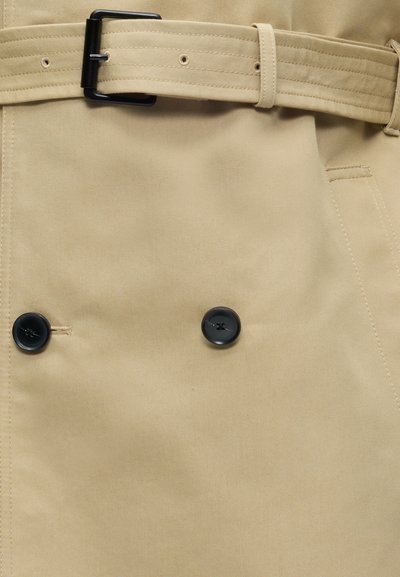 Tissu beige pour trench-coat avec des boutons noirs et une ceinture assortie en tissu avec une boucle rectangulaire noire.