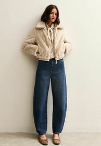 Chaqueta bomber de piel sintética beige sobre un suéter crema, combinada con jeans azul oscuro de tiro alto y pierna ancha, y zapatos planos marrón claro.