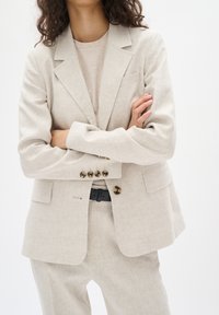 Beige linneblazer med standard slag, fyra knappkn�äppningar och sidfickor. Matchas med en matchande beiga outfit. Slät textur.