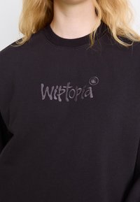 Sweat-shirt noir avec le mot « Wistopia » brodé en fil foncé sur la poitrine, porté par une personne aux cheveux blonds.