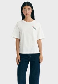 Junge Frau mit langen schwarzen Haaren trägt ein weißes übergroßes T-Shirt mit dem Aufdruck "Thinking Mind" und einer Blumen-Grafik, kombiniert mit dunkelblauen Jeans.