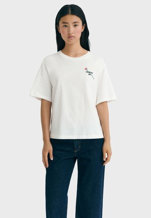 Jonge vrouw met lang zwart haar draagt een wit oversized T-shirt met "Thinking Mind" en een bloemenafbeelding, gecombineerd met donkerblauwe jeans.