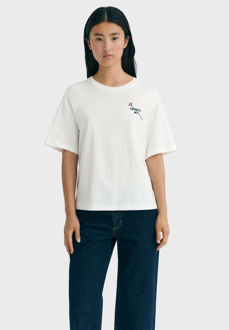 Junge Frau mit langen schwarzen Haaren trägt ein weißes übergroßes T-Shirt mit dem Aufdruck "Thinking Mind" und einer Blumen-Grafik, kombiniert mit dunkelblauen Jeans.