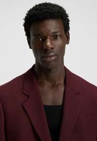 Burgundy blazer with notch lapels, textured fabric, and a single chest pocket. Worn over a black tank top. Close-up portrait.