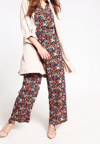 Mono floral en negro con flores de colores; tejido ligero, piernas anchas, cierre delantero con botones, combinado con una chaqueta ligera beige.
