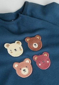 Mörkblått tyg med fyra broderade björnhuvuden i kräm, brun, ljusbrun och mörkrosa ordnade i ett rutmönster.
