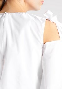 Biała bluzka z odkrytymi ramionami, wyróżniająca się wiązanym akcentem na ramieniu. Gładka tekstura materiału z dyskretnym zapięciem na zamek z tyłu.
