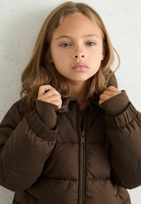 Brūns pūkains jaka ar rāvējslēdzēju priekšpusē, elastīgām aprocēm un kapuci. Audums ir gluds ar polsterētu daļu, lai nodrošinātu siltumu.