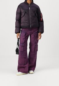 Pěnová černá bomber bunda se zipovým zapínáním, kombinovaná s širokými fialovými cargo kalhotami s několika kapsami a strukturovanou tkaninou.