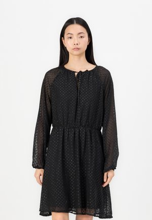 Black dress made of sheer fabric with a textured polka dot pattern, long sleeves, elastic waist, and tie detail at the neckline.