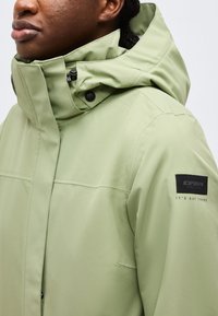 Personne portant une veste à capuche vert clair avec un col haut et un écusson noir sur le haut du bras indiquant « ICEPEAK IT'S OUT THERE ».