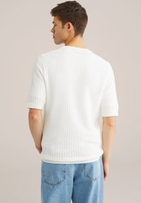 Jonge man met de rug naar de camera, gekleed in een witte geribbelde korte mouw trui en lichtblauwe spijkerbroek, tegen een effen achtergrond.