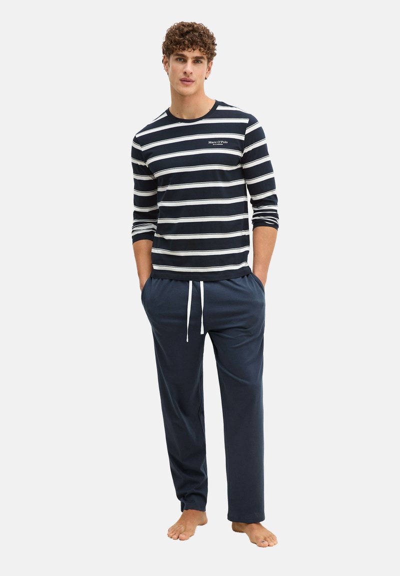 Man draagt een navyblauw-wit gestreept shirt met lange mouwen en donkerblauwe broek met trekkoord, staat op blote voeten met handen in de zakken.