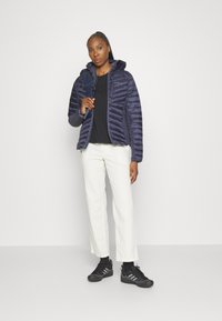 Marineblauwe pufferjack met een getailleerd ontwerp, rits aan de voorkant en capuchon; gedragen over een zwart shirt en gecombineerd met witte broek en zwarte sneakers.