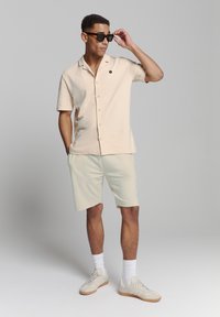 Beige korte mouw boutonshirt met kraag; gecombineerd met lichte shorts; model draagt zwarte zonnebril; schoenen in neutrale tinten.