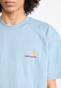 Light blue cotton t-shirt featuring a yellow embroidered logo and red text on the left chest. Classic crew neckline and short sleeves.