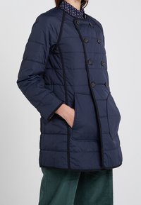 Tummansininen toppatakki, jossa on kaksirivinen malli, suuret napit, kaksi taskua ja kontrastina mustat reunukset saumoissa.