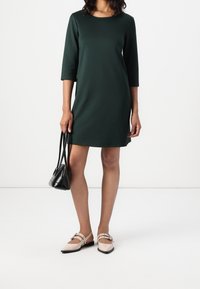 Nainen, joka pukeutunut tummanvihreään polvipituiseen mekkoon, pitää pientä mustaa käsilaukkua, ja sen parina hänellä on beigeitä korkokenkiä, joissa on soljet.