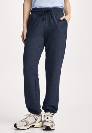 Person trägt marineblaue Jogginghose mit elastischen Bündchen und weiße Sportschuhe, steht vor hellem Hintergrund.