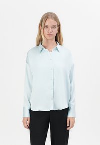 Lyseblå, langermet skjorte med knapper og klassisk krage, glatt tekstur og en litt løs passform, båret med svarte bukser.