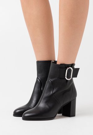 Bottes chevilles en cuir noir avec une surface texturée, un bout pointu, un talon carré et un détail de boucle sur la sangle de la cheville.