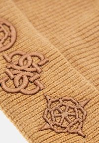 Berretto lavorato a maglia in un caldo colore beige, caratterizzato da ricami tono su tono con disegni intricati. Texture a coste distintive su tutta la superficie.