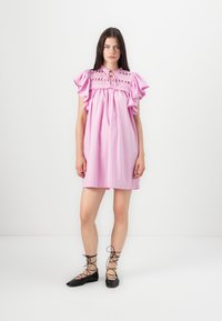 Rozā kokvilnas kleita ar viļņainām piedurknēm un izšūtu jostu. Pāris melnu šņorējamu apavu, stāvot uz balta fona.