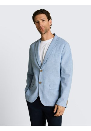 Man draagt lichtblauwe blazer over wit overhemd en donkere broek, staand tegen een egale lichte achtergrond.