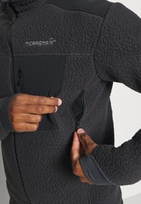 Giacca in pile nera con pannelli spalla lisci neri. Presenta tasche con zip e aperture per il pollice sui polsini per un comfort maggiore.