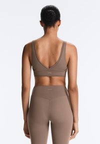 Woman seen from the back wearing a matching taupe sports bra and high-waisted leggings with hair in a bun against a plain background.