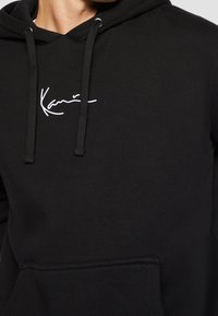 Gros plan sur une personne portant un sweat à capuche noir avec le logo "Karrimor" en écriture cursive blanche, des cordons noirs et une poche kangourou à l'avant.