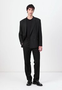 Black suit comprising a single-breasted jacket with lapels and trousers, paired with a black shirt and shiny black shoes on a neutral background.