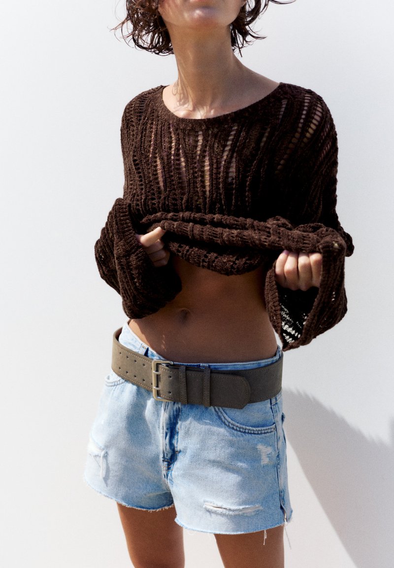 Personne soulevant un pull en maille marron, portant un short en jean bleu clair avec une grande ceinture grise, sur fond blanc uni.