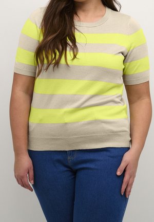 Vrouw draagt een beige trui met korte mouwen met felle gele horizontale strepen en een blauwe spijkerbroek, staand tegen een lichte achtergrond.