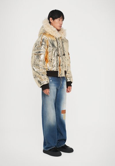 Gouden metallic pufferjack met nepbont kraag, zwarte geribbelde manchetten en een ritssluiting aan de voorkant. Draagt een lichtblauwe wijde jeans met patches.