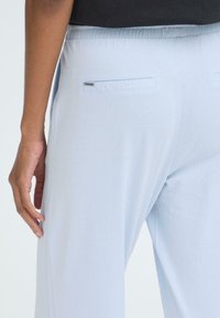 Lyseblå bukser lavet af blødt stof med en glat tekstur, elastisk taljebånd og to baglommer med små lukninger.