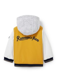 Żółta i biała kurtka przeciwwiatrowa z szarym kapturem, ribbowanymi mankietami oraz napisem "Zespół szopów" na plecach.
