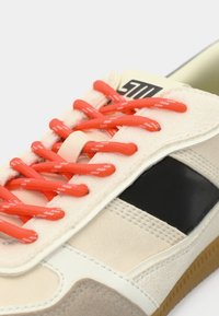 Baskets beiges avec bande noire, lacets rouges, accents blancs et semelle en caoutchouc texturée en gros plan.